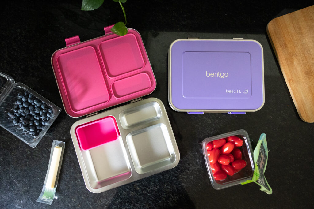 Open Bentgo Kids stainless steel lunch boxes in pink and purple with three-compartment trays, surrounded by blueberries, string cheese, and grape tomatoes on a black kitchen countertopOpen Bentgo Kids stainless steel lunch boxes in pink and purple with three-compartment trays, surrounded by blueberries, string cheese, and grape tomatoes, Raleigh NC documentary family photography