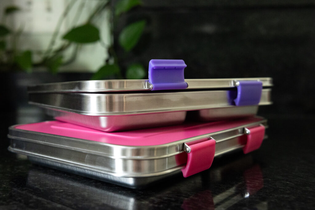 Stacked stainless steel Bentgo Kids lunch boxes with purple and pink lids on black granite countertop, photographed by Raleigh NC family photographer. One purple latch is flipped up and others are closed, showcasing how easy it is to open and close them around the stainless steel rim around the side.