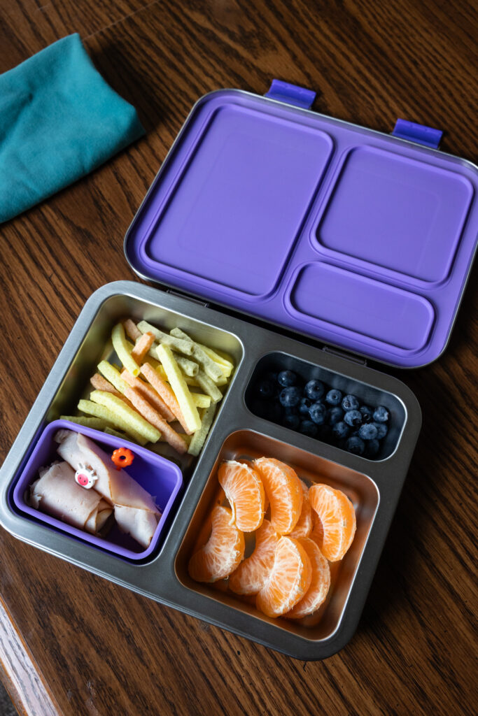A purple-lidded stainless steel Bentgo kids lunch box sits open on a wooden table, showing its kids lunch contents of veggie straws, meat & cheese roll-ups with plastic toothpicks, orange slices, and blueberries, photographed by Raleigh NC family photographer Light Creative