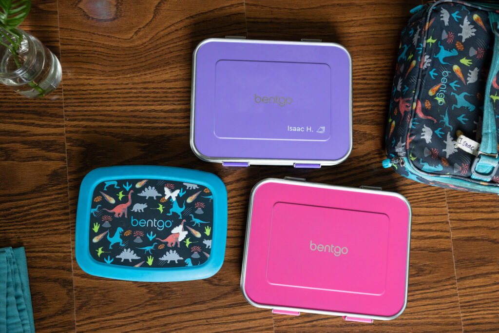 An aerial view of three Bentgo kids lunch boxes sitting closed on a wooden table, two stainless steel with purple and pink lids and one dinosaur print plastic Bentgo kids prints lunch box, next to a matching dinosaur print Bentgo kids lunch bag. All photographed by family product photographer Light Creative in Raleigh NC